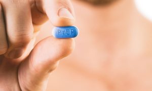 A man holds a blue PrEP pill between his fingers, face blurred in background