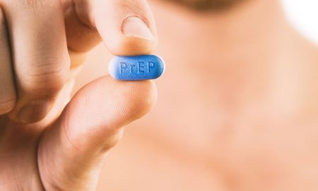A man holds a blue PrEP pill between his fingers, face blurred in background