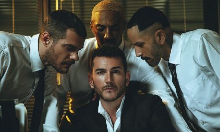 Jim Verraros in the Good Boy music video, seated in a suit surrounded by men in white shirts