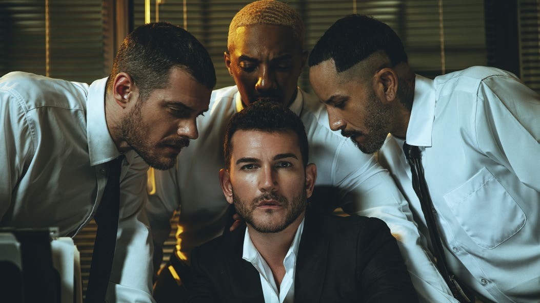 Jim Verraros in the Good Boy music video, seated in a suit surrounded by men in white shirts